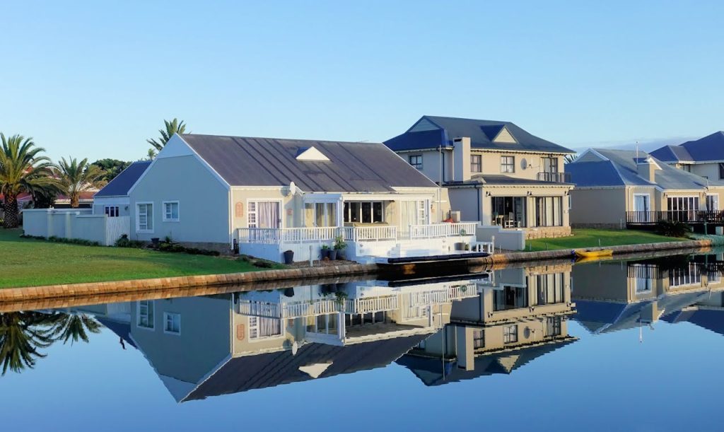 Luxury modern homes reflecting on a peaceful, clear lake during daylight, showcasing architectural beauty.